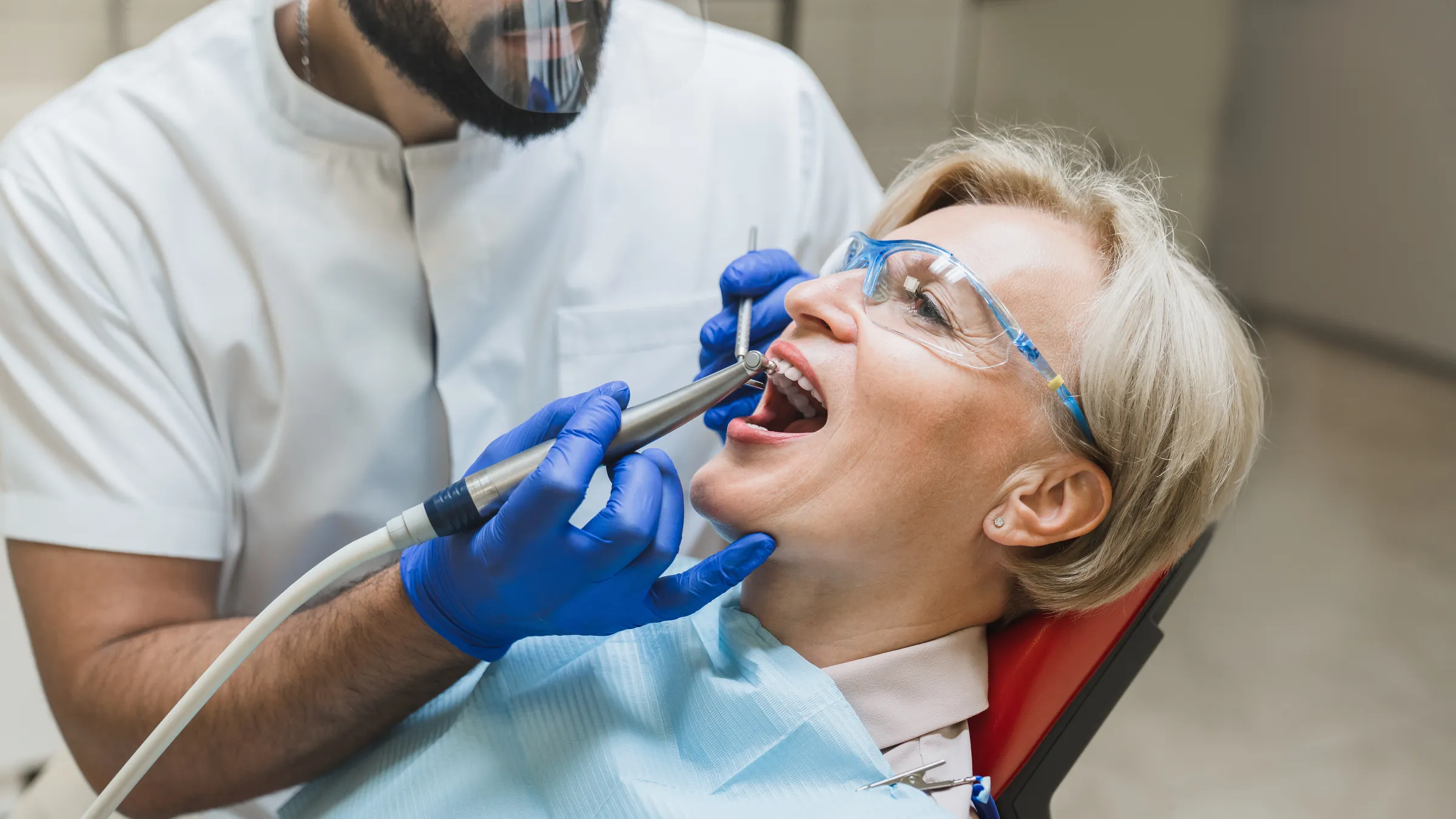 A dentist in a face shield and gloves preparing a patient's upper tooth with a handpiece