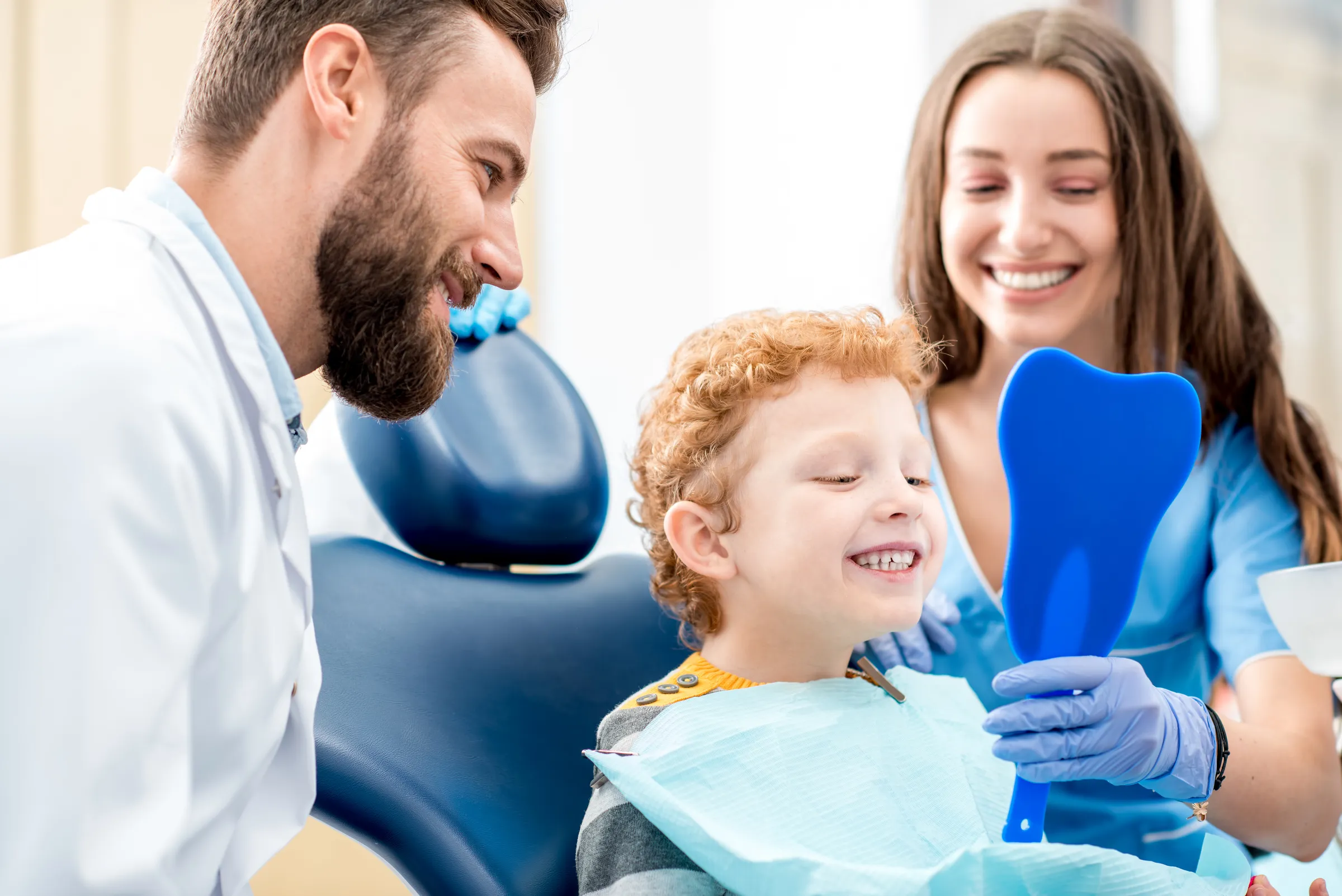 A dentist and a parent beside a young child holding a mirror in the dental chair