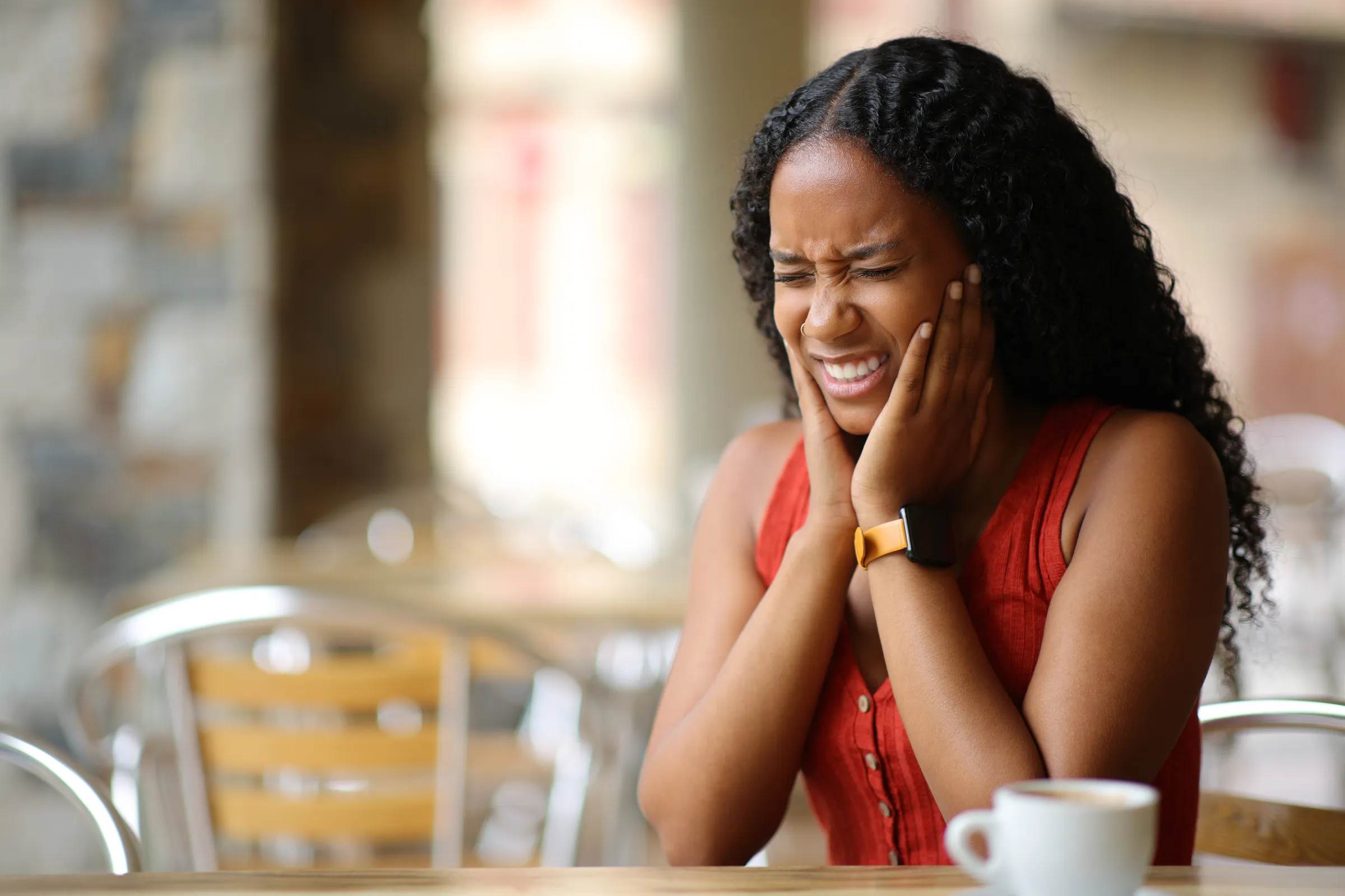 A woman holding her jaw in discomfort while sitting at a cafe table